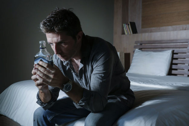 Man battling alcohol addiction sitting on his bed with bottle of alcohol
